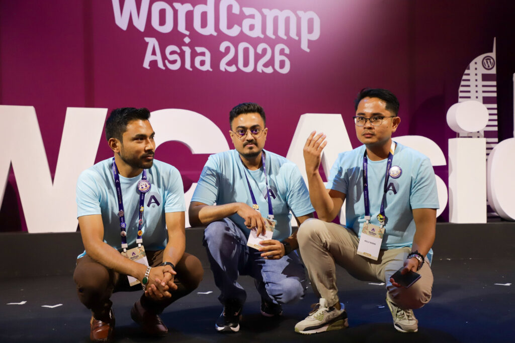 Rolly Bueno crouching on stage with two fellow WordCamp Asia 2026 Contributor Day organizers in front of the event backdrop.
