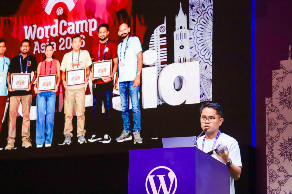 Rolly Bueno speaking at a podium on stage during WordCamp Asia 2026, with a presentation slide displayed behind him.